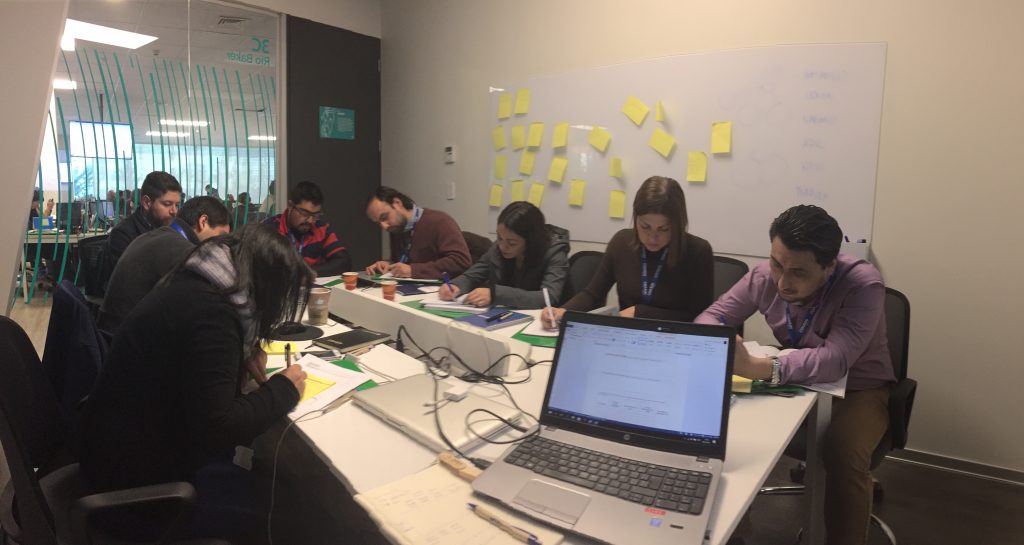 Personas sentadas alrededor de una mesa de conferencias, trabajando con documentos. Hay una computadora portátil abierta sobre la mesa y, al fondo, una pizarra con notas adhesivas.