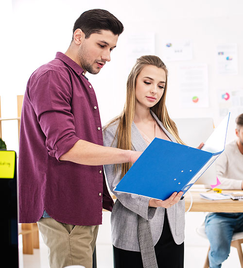Dos personas de pie y mirando una carpeta azul juntas en un entorno de oficina, con papeles y una computadora visibles en el fondo.